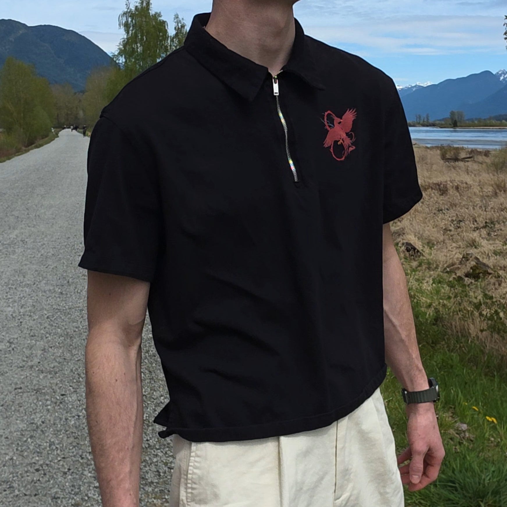 Man wearing a black polo shirt with a red logo and beige pants walking on a road with mountains in the background.