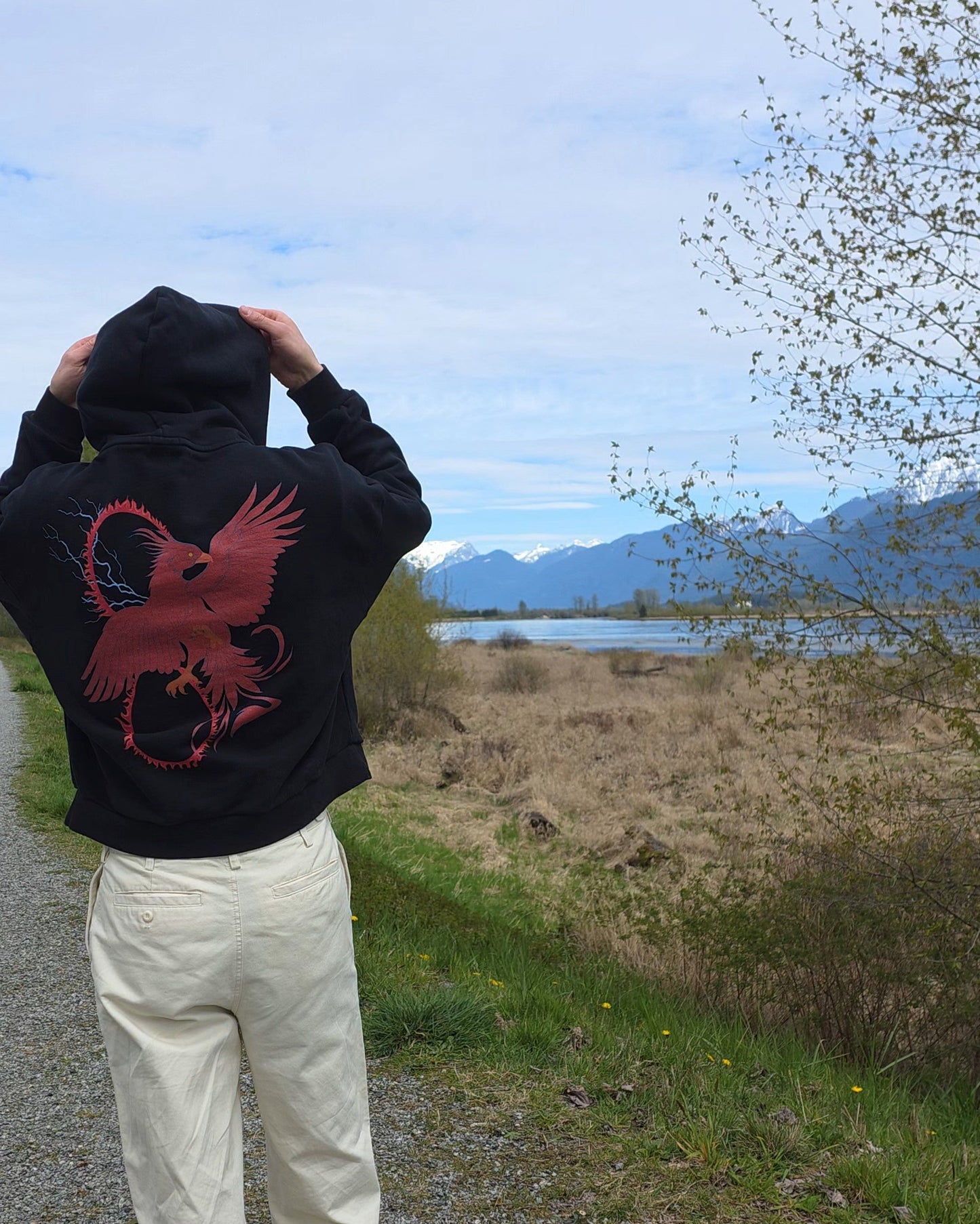 Person wearing a black hoodie with a RISE red pheonix design, standing on a path with a scenic background of mountains and water.