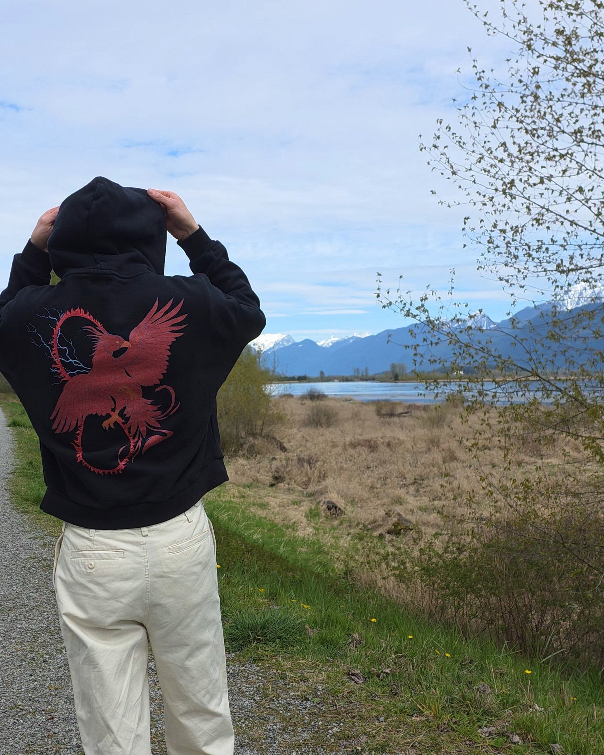 Person wearing a black hoodie with a RISE red pheonix design, standing on a path with a scenic background of mountains and water.