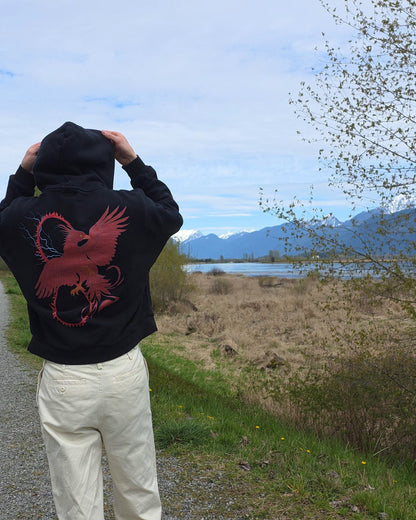 Person wearing a black hoodie with a RISE red pheonix design, standing on a path with a scenic background of mountains and water.