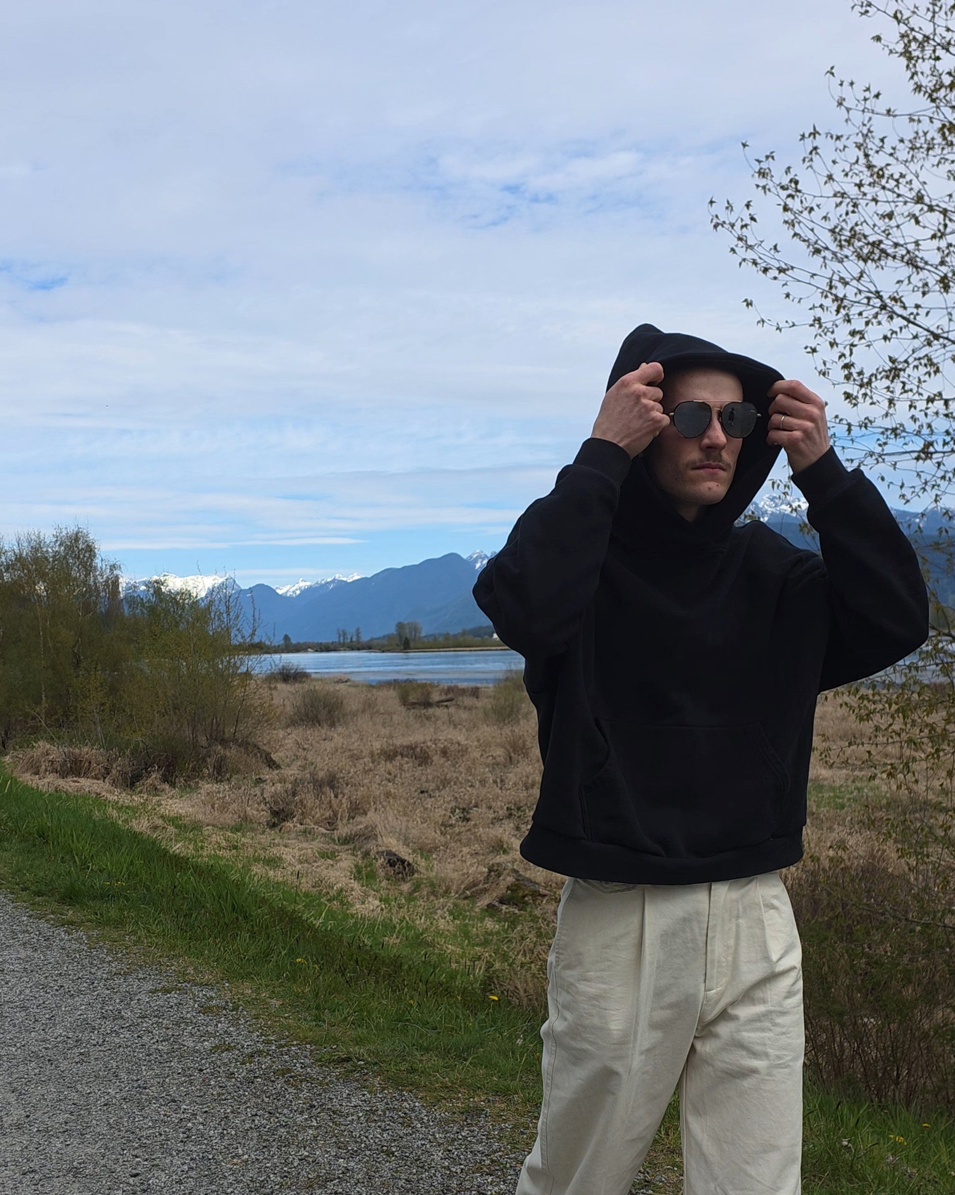 Person wearing a black RISE Oversized hoodie and white FUNDAMENTAL Straight Slacks standing on a path with a scenic background.