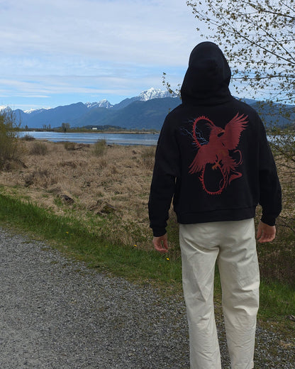 Person wearing a black RISE Oversized hoodie with a red pheonix design, standing on a path with mountains and a lake in the background.
