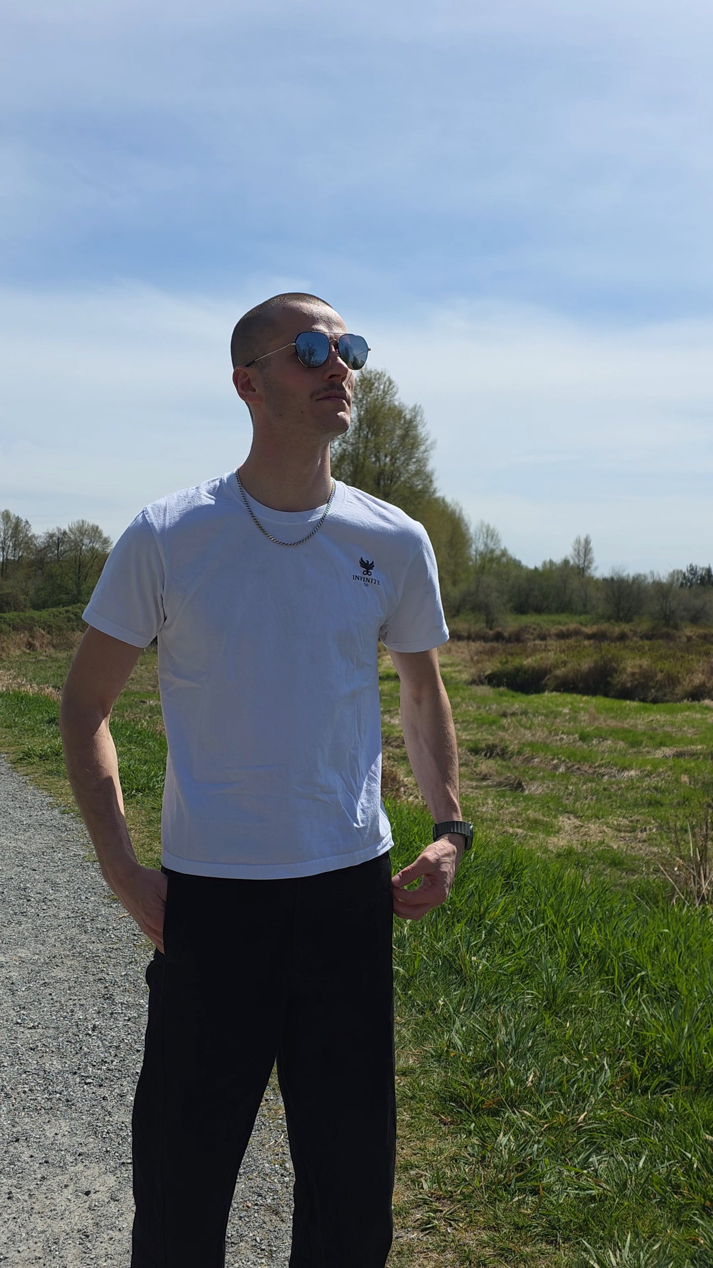 Man wearing sunglasses and a white t-shirt with a Infinite Co logo standing on a path with grass and trees in the background.