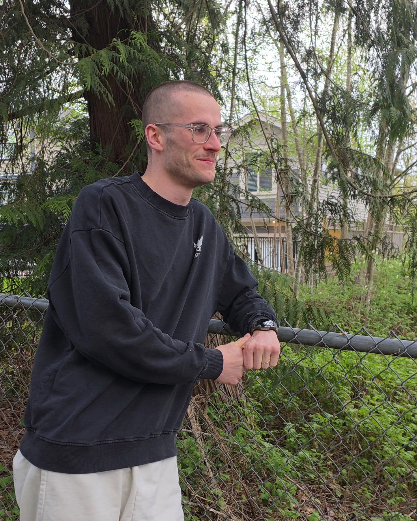 Man standing outdoors wearing an EDEN Oversized Crewneck, holding onto a fence with trees and a house in the background