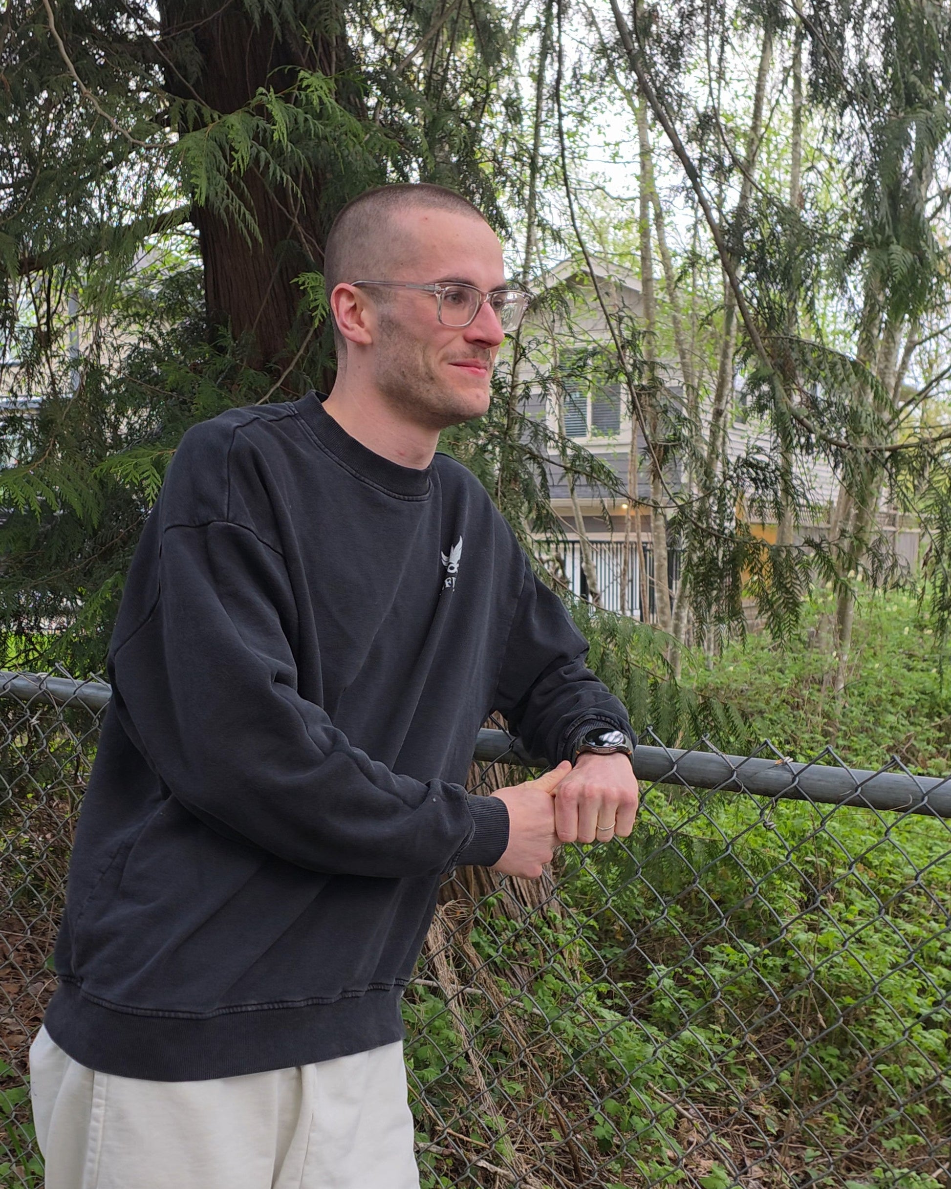 Man standing outdoors wearing an EDEN Oversized Crewneck, holding onto a fence with trees and a house in the background